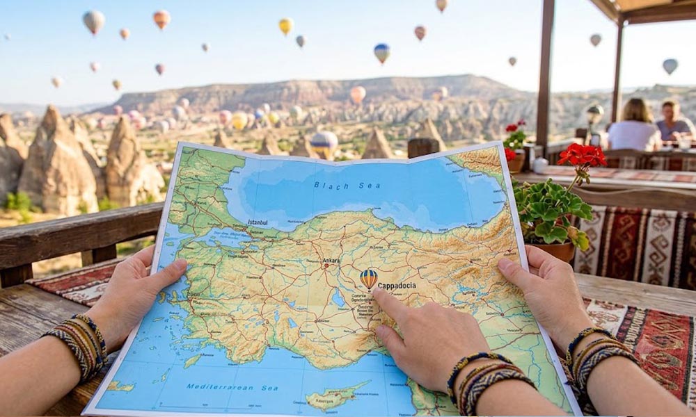 Kadar iz prvog lica na ruke sa šarenim narukvicama koje drže raširenu geografsku mapu Turske i Kapadokije na drvenom stolu na terasi. U pozadini se vide karakteristične stenske formacije Kapadokije i šareni baloni na topao vazduh na vedrom plavom nebu. Slika je svetla i živopisna.
